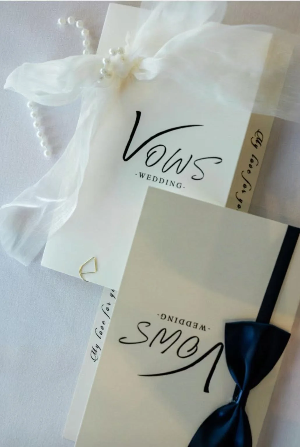 Wedding invitation with a white ribbon bow and a dark blue satin bow, featuring black and gold lettering.