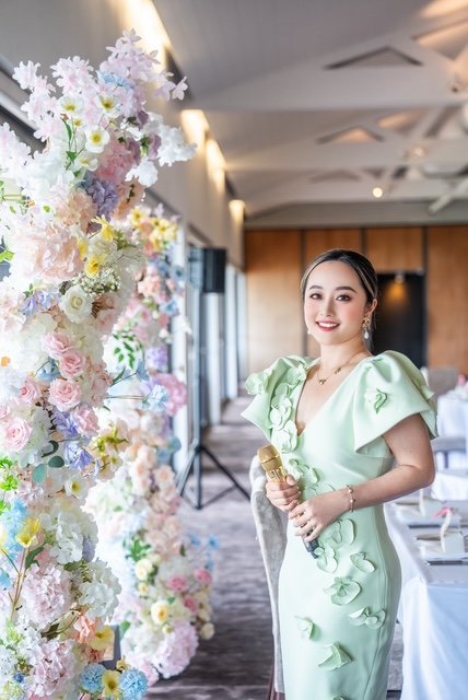 A woman in a light green dress standing beside a large floral arrangement in an indoor event space.