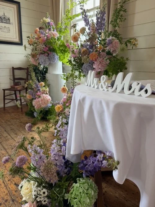 Table with 'Mr. & Mrs.' sign, surrounded by colorful flower arrangements, in a decorated indoor setting.