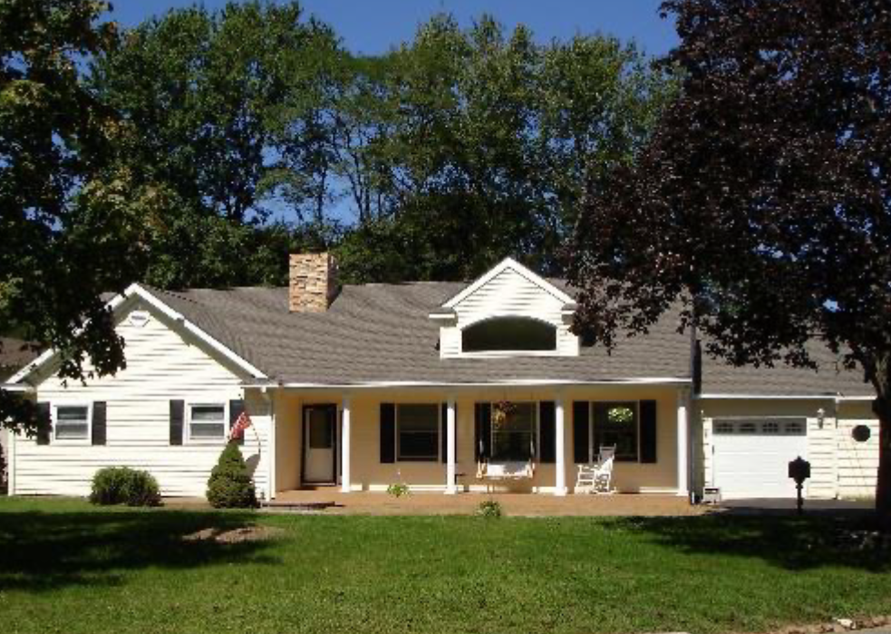 A white house with black shutters and a front porch, surrounded by green grass and trees, with a cloudy sky in the background.