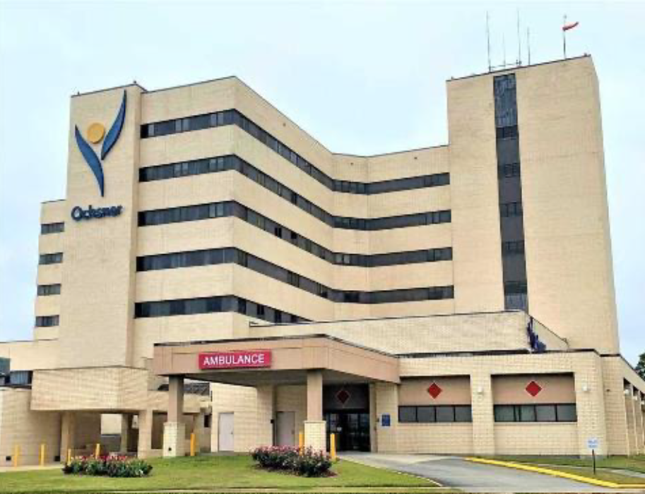 Hospital building with a sign for ambulance, a logo on the top left, and a small garden in front.