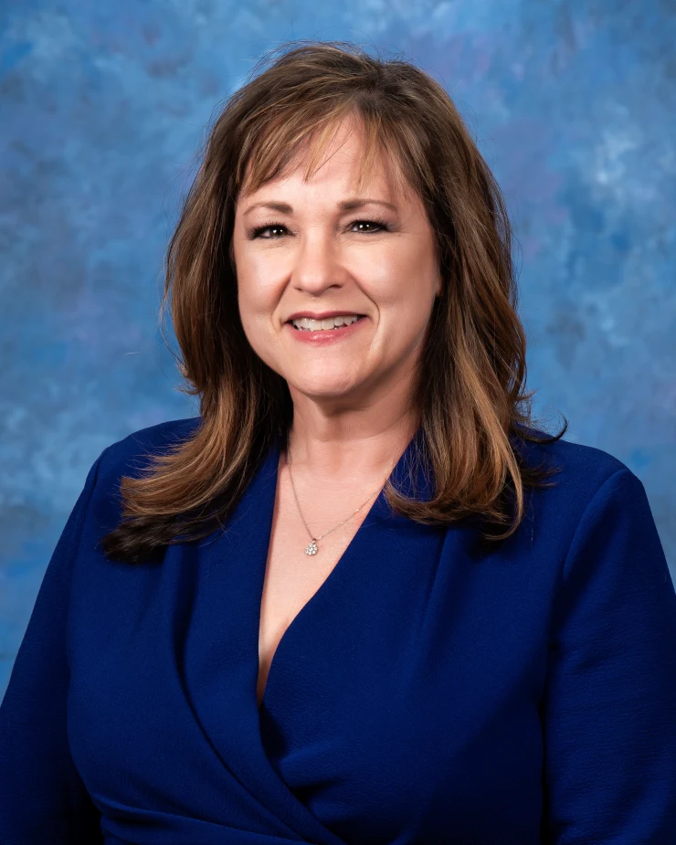 A smiling woman with brown hair wearing a blue blazer and a delicate necklace against a blue patterned background.