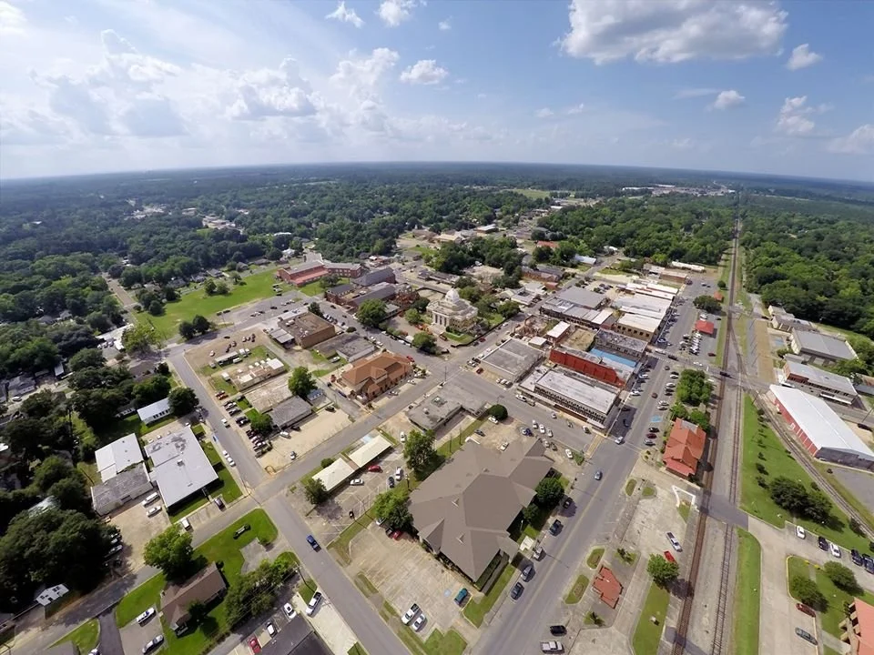 Aerial view of a small town with a church, commercial buildings, parking lots, and surrounding greenery under a partly cloudy sky.