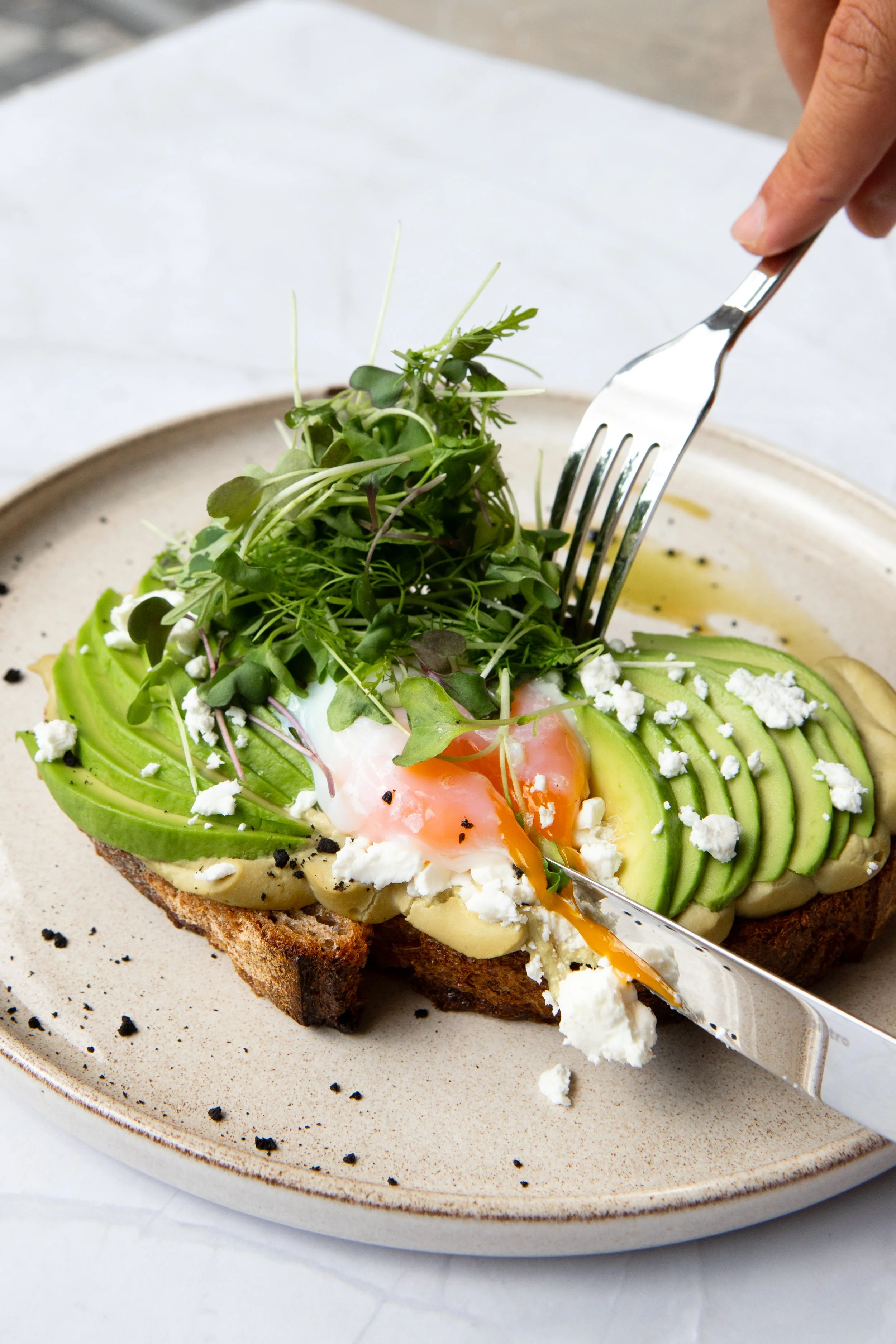 Open-faced avocado toast with microgreens, soft-poached egg, crumbled cheese, and black pepper on a toasted bread slice, with a fork cutting into it.