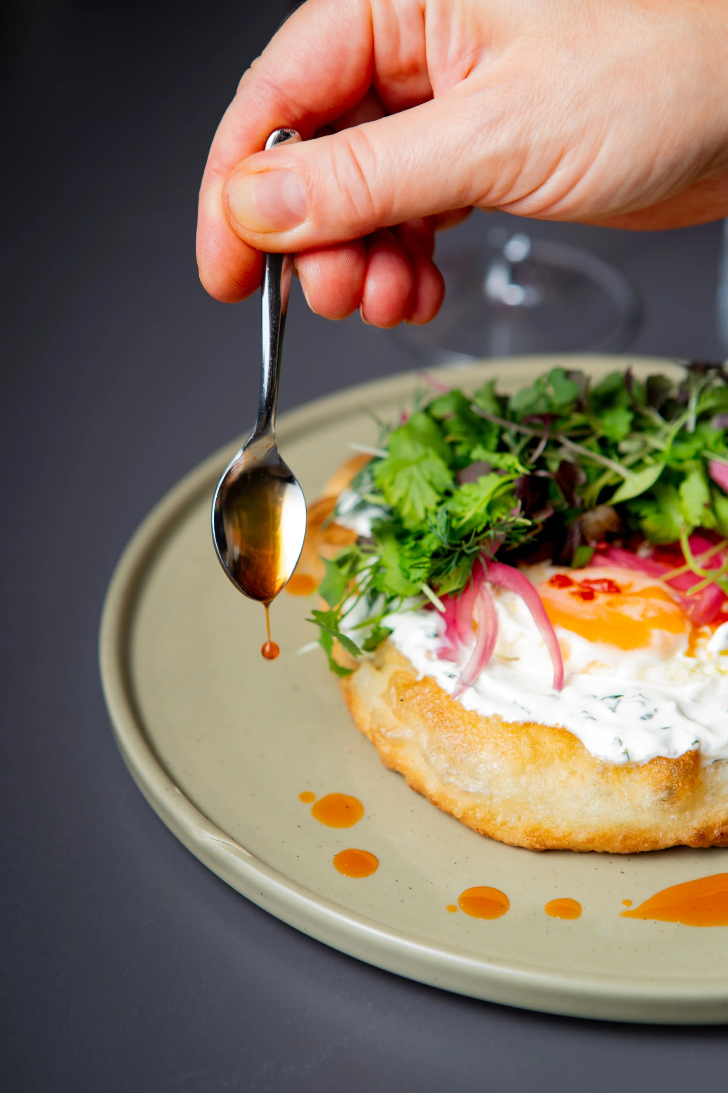 A person drizzling sauce over a dish with fried egg, salad greens, and vegetables on a beige plate.