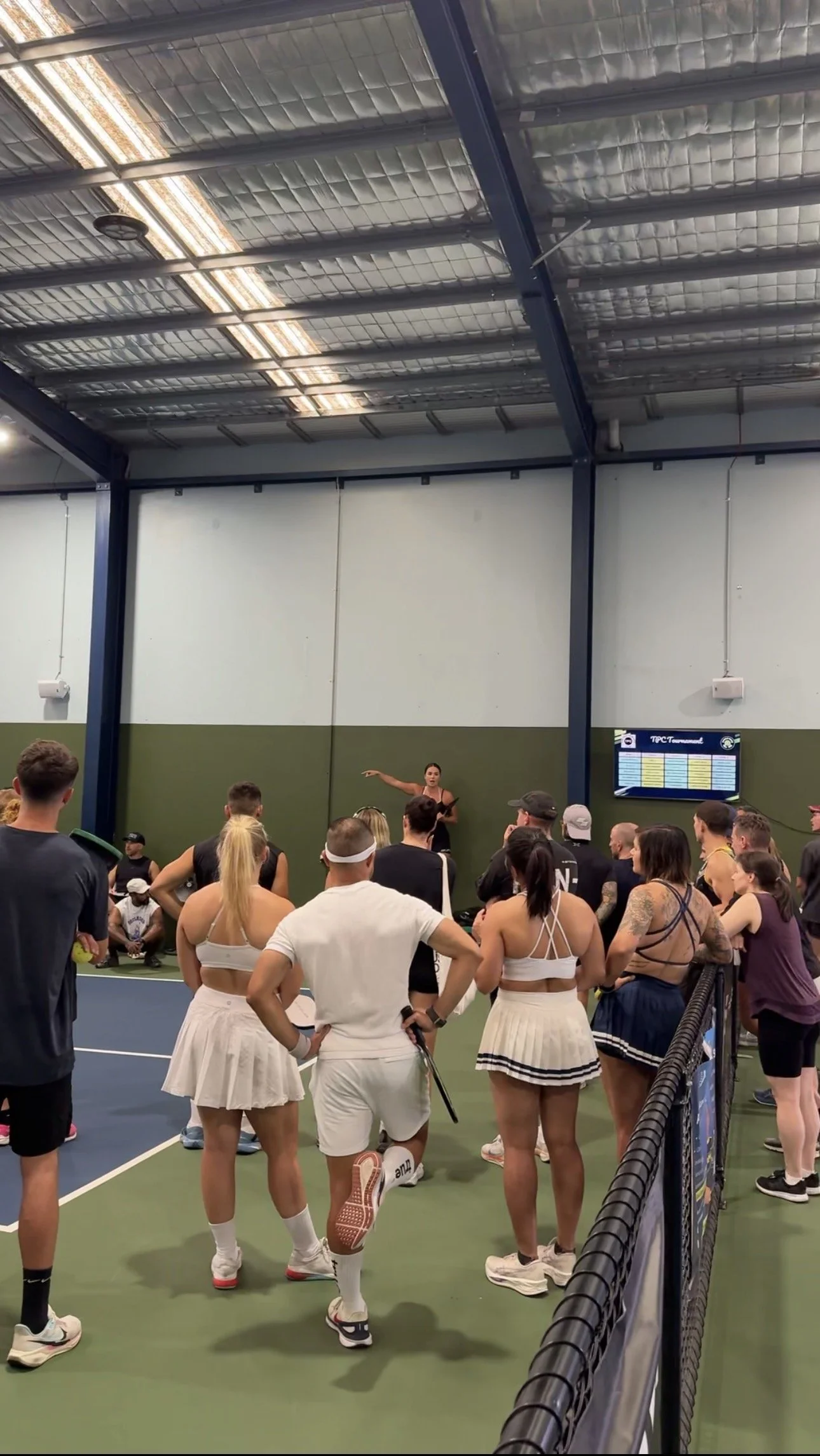 A group of tennis players listening to a female coach giving instructions on an indoor tennis court.