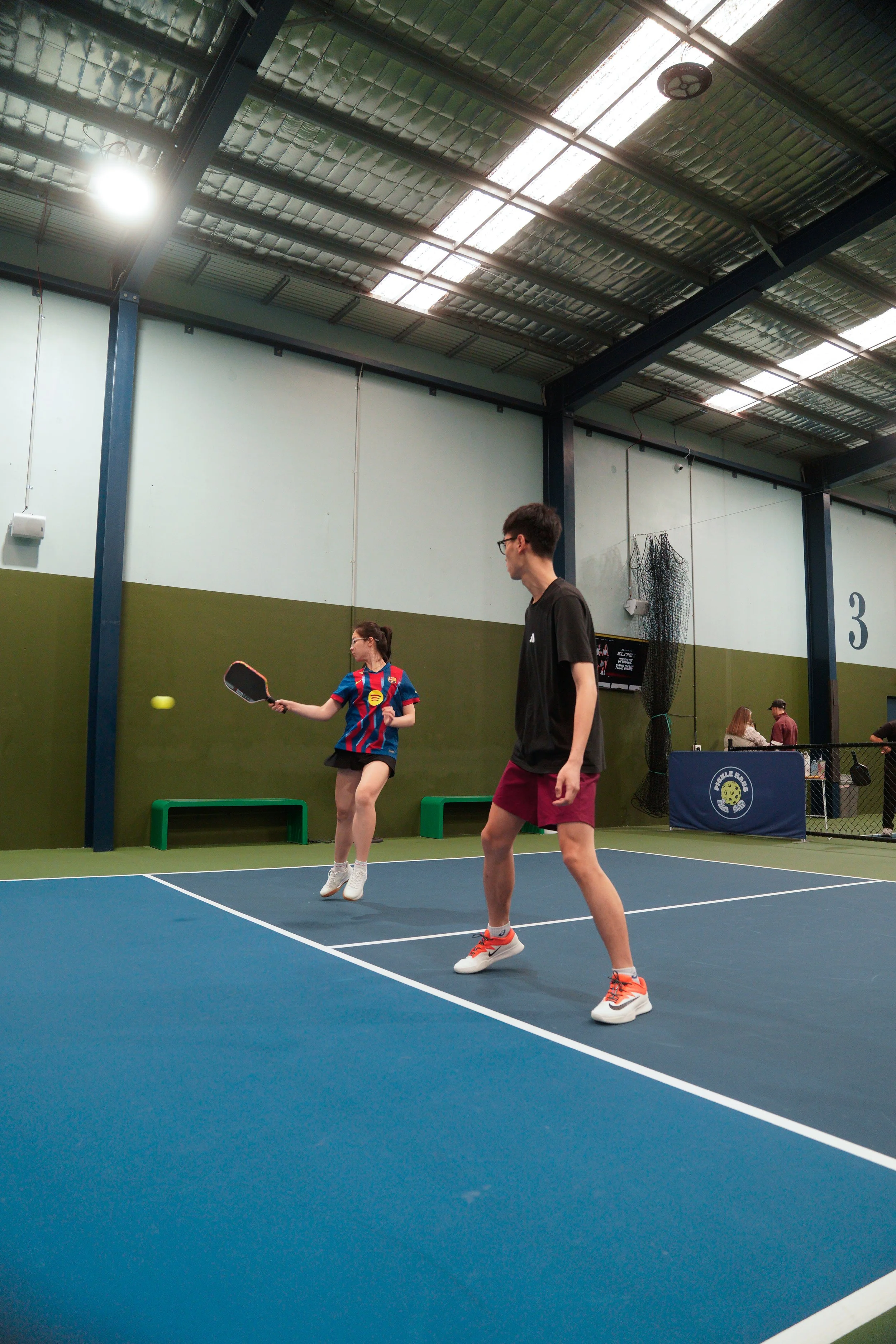 Two people playing pickleball on an indoor court, one woman hitting the ball with a paddle and one man standing nearby, with people watching in the background.