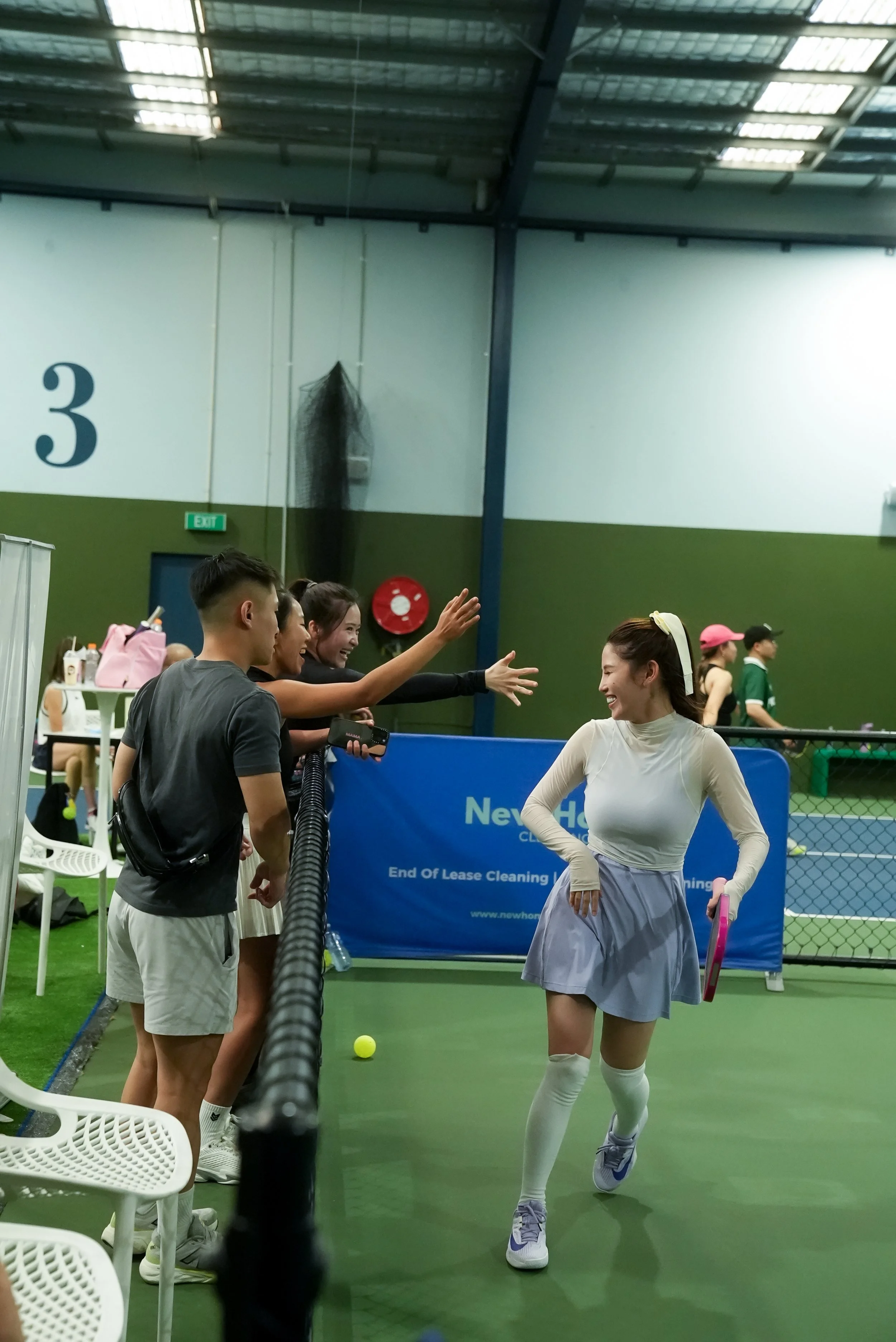 A woman in a tennis outfit walking on an indoor tennis court while smiling, with friends cheering her on from behind a black fence.