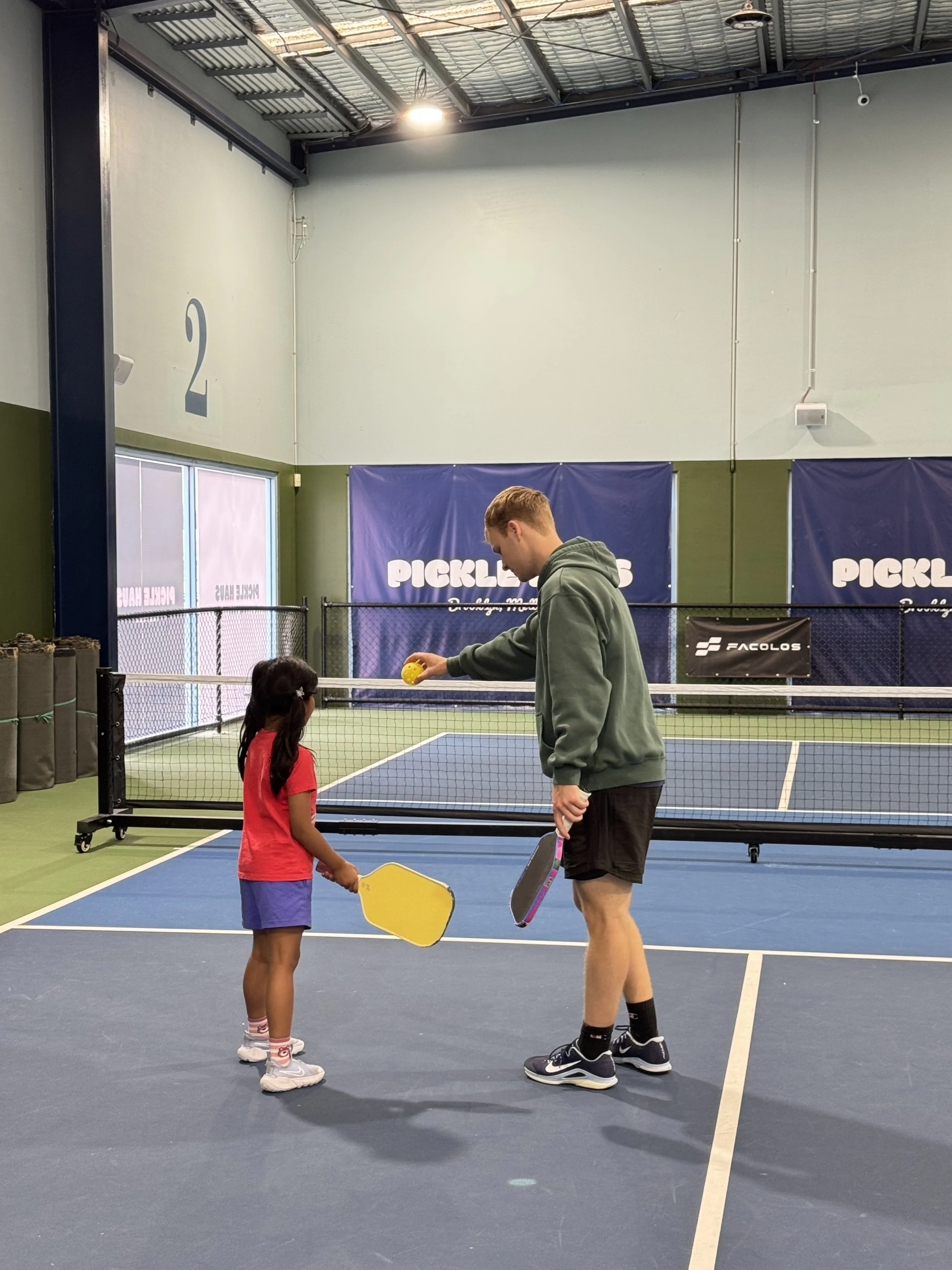 A young girl and a man playing pickleball indoors on a court, with the girl holding a yellow paddle and the man holding a paddle and a pickleball in his hand.