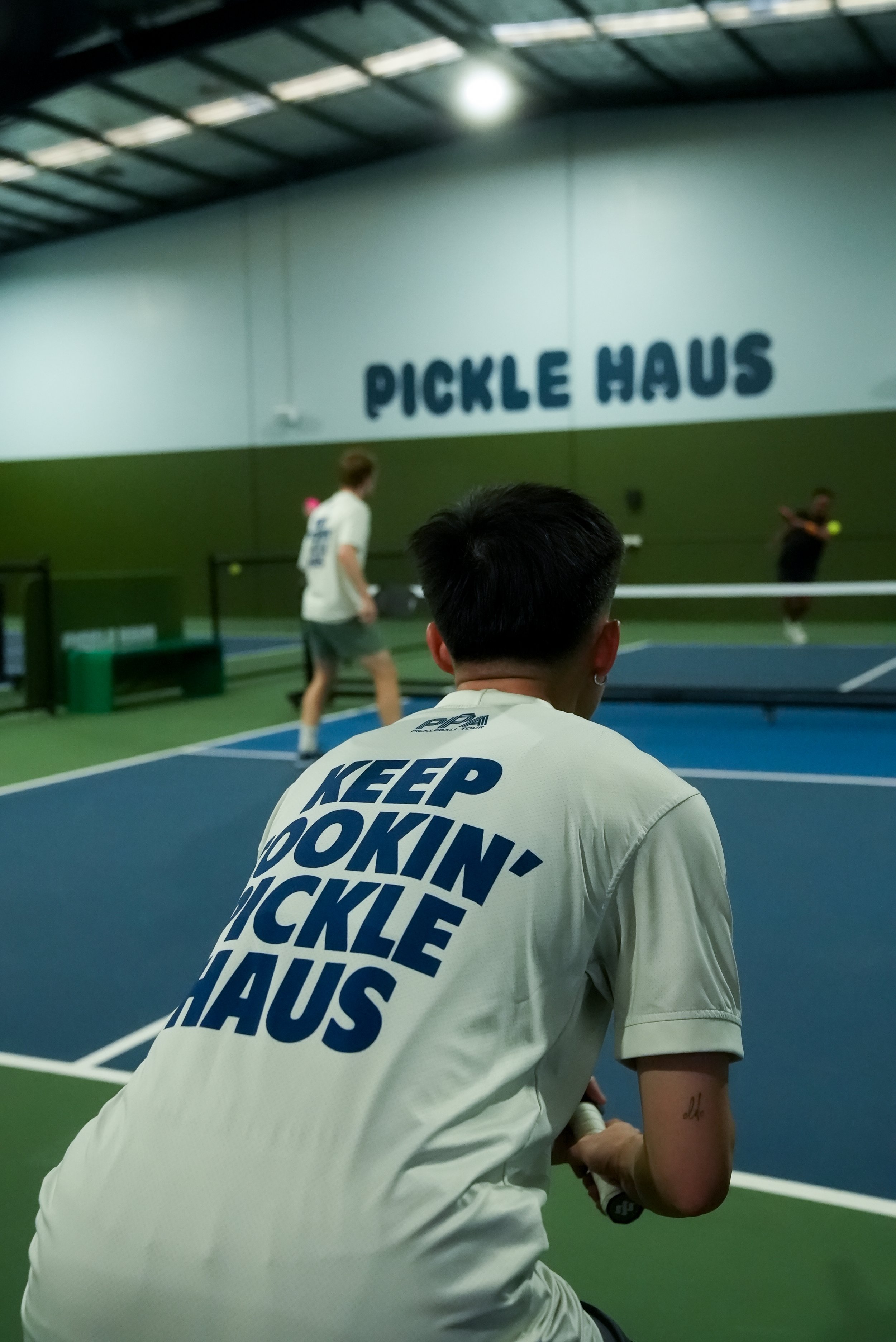 People playing pickleball at Pickle Haus gym with a large sign on the wall that says "PICKLE HAUS." One person in the foreground wearing a cream-colored shirt with "KEEP LOOKIN' PICKLE HAUS" written on the back.