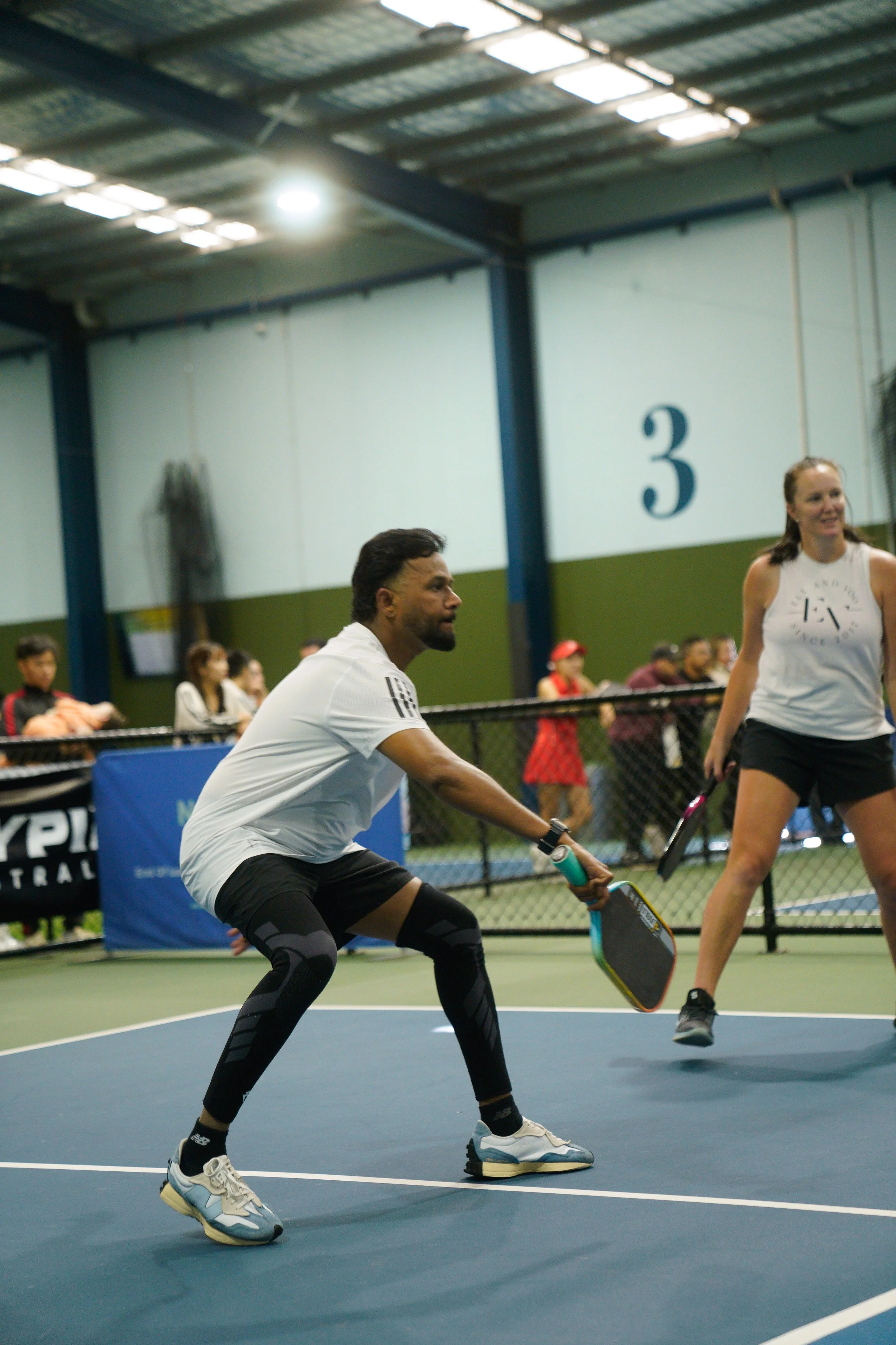 A man and woman playing pickleball on an indoor court, with onlookers behind a black fence.