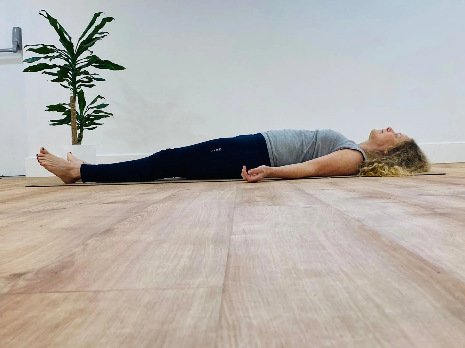 A woman lying on her back on a yoga mat, practicing relaxation or meditation, with a potted plant in the background.