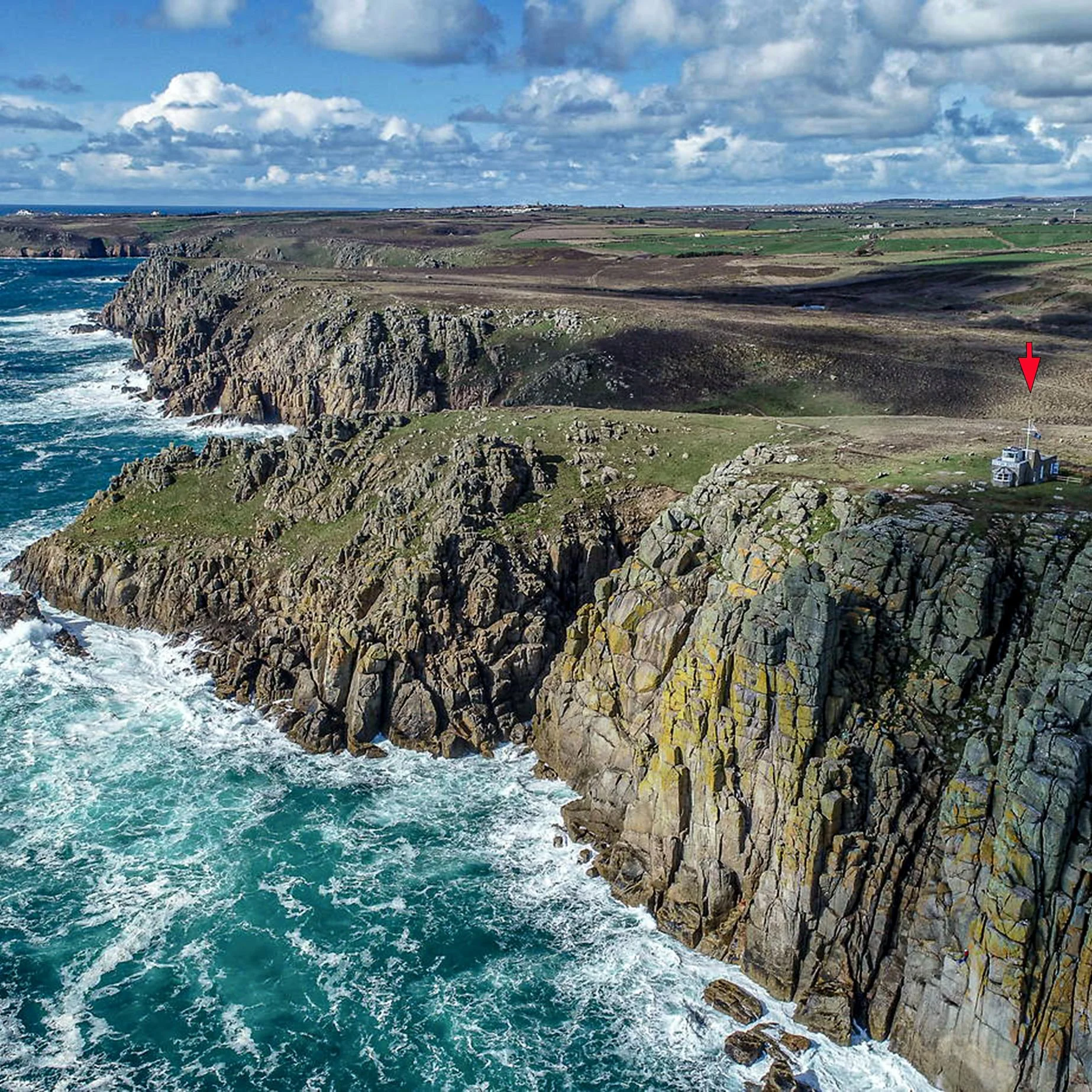 Cliffs overlooking the ocean with rocks and waves, green fields on top, and a house with an American flag in the distance.