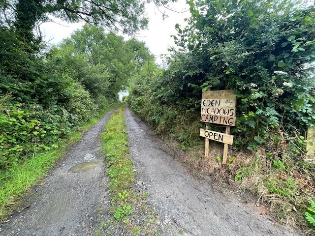 Gravel rural road leading into a wooded area, with a handwritten sign on the right side that reads 'Eden Meadows Camping Open'