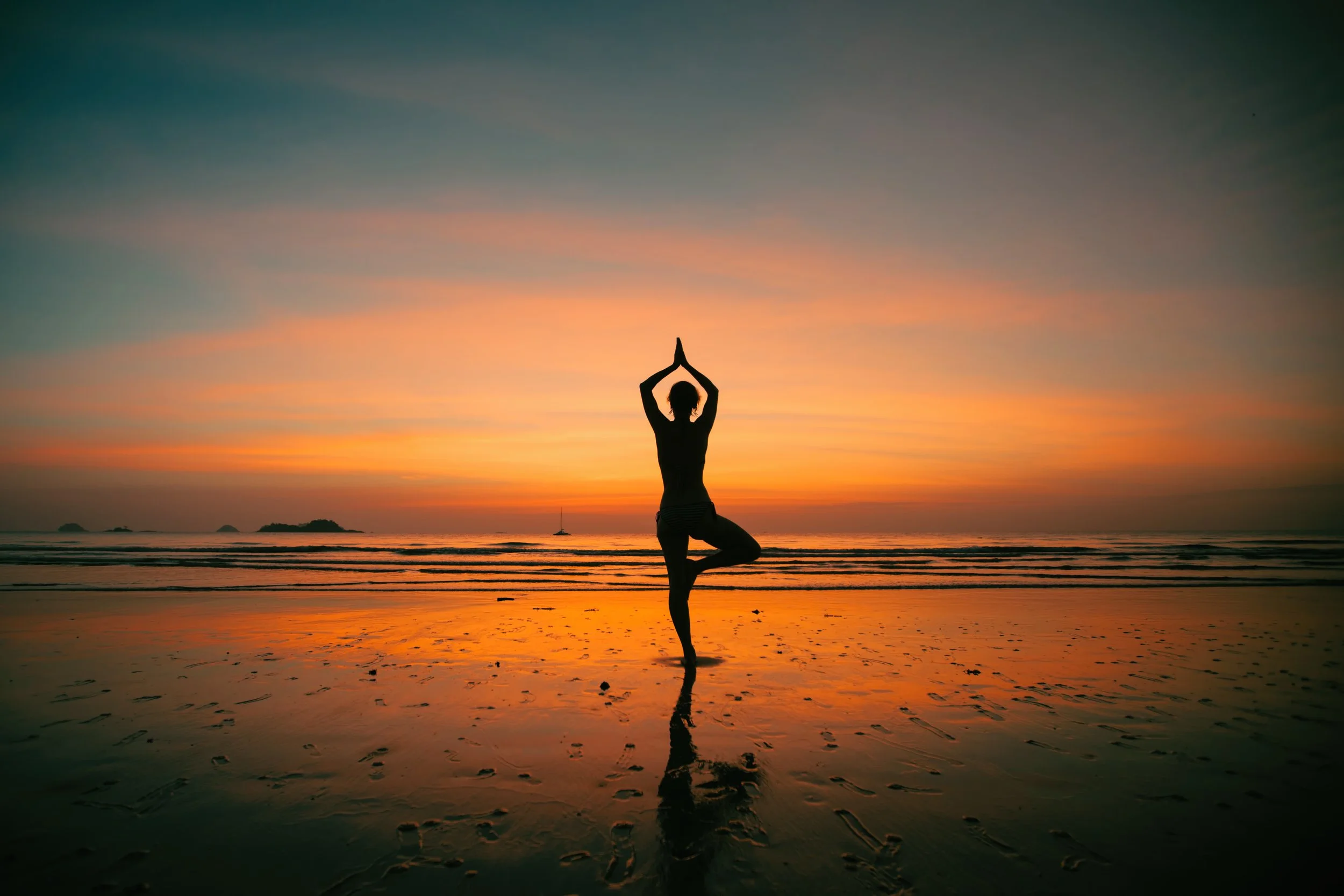 Person practicing yoga in tree pose on a beach during sunset with an orange and purple sky.
