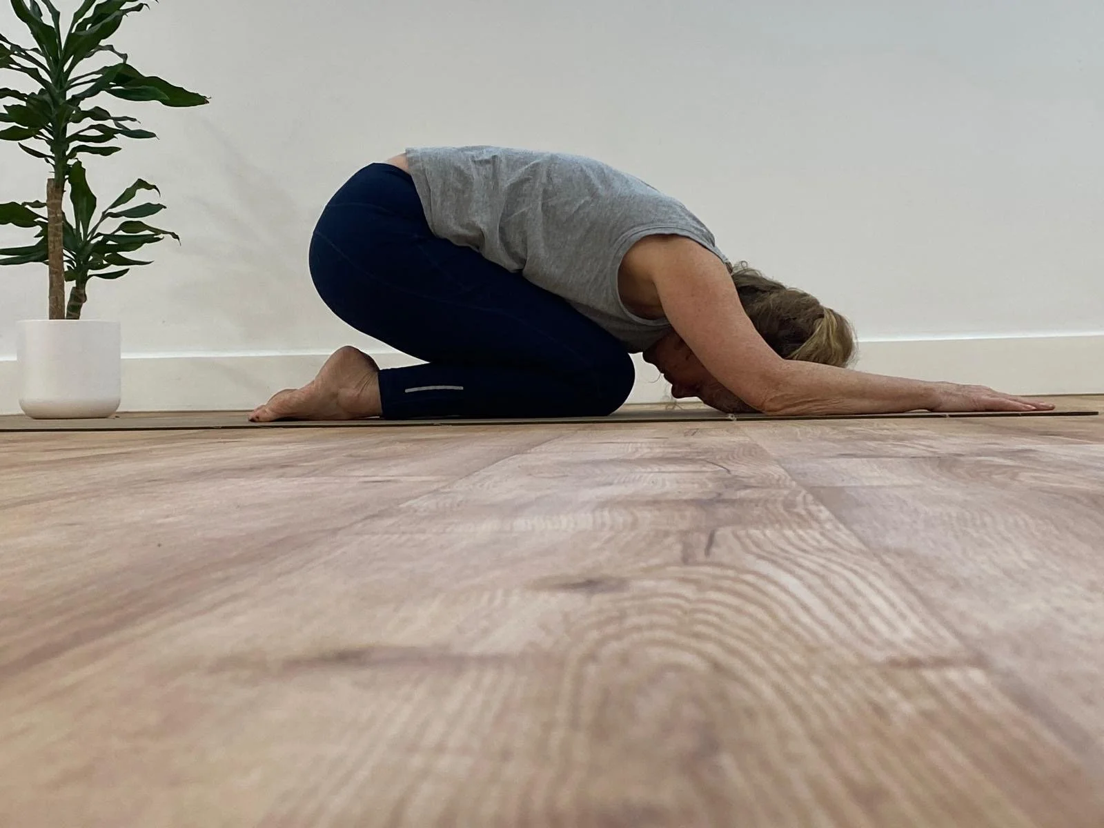 A woman in a yoga pose called child’s pose on a wooden floor, with a potted plant nearby in a minimalistic room.