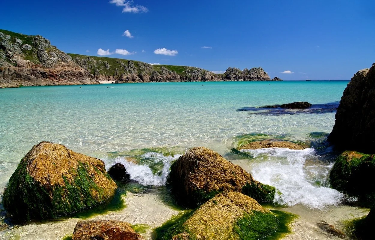 A scenic beach with clear turquoise water, green moss-covered rocks in the foreground, rocky cliffs on the left, and a bright blue sky with a few white clouds.