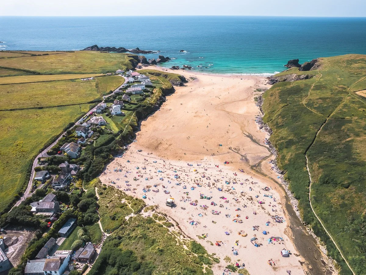 Aerial view of a sandy beach with colorful umbrellas, surrounded by green grassy hills and a row of houses on the left side near the coast.