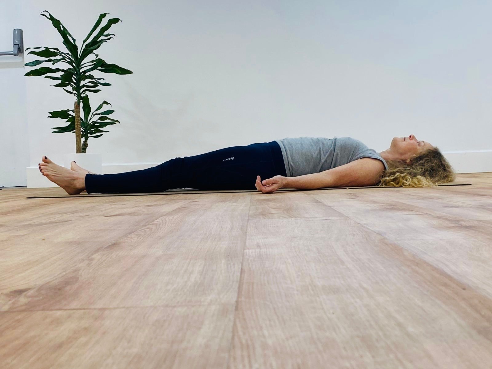 A woman lying on her back on a yoga mat in a peaceful indoor setting, with a white wall and a potted plant nearby.