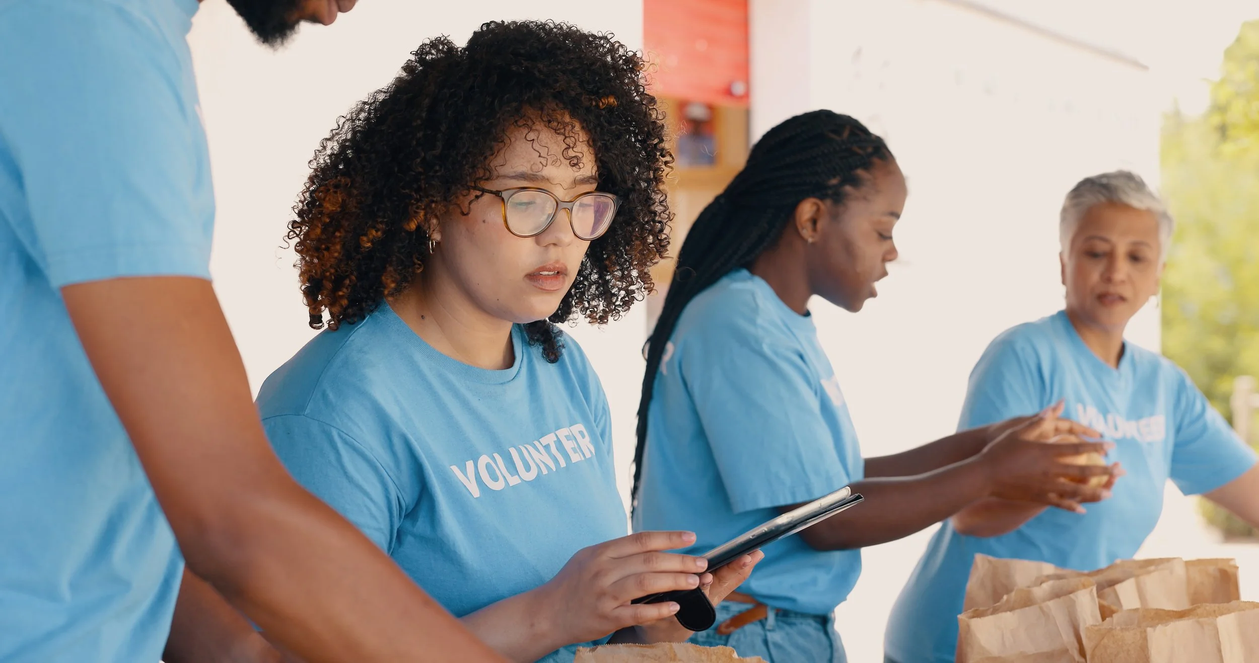 Group of volunteers wearing blue shirts with 'VOLUNTEER' printed on them, working together at an outdoor event, preparing food or supplies, with trees in the background.