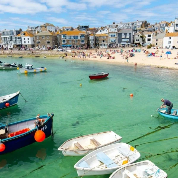 Boats anchored near a sandy beach with colorful houses and people sunbathing and swimming.