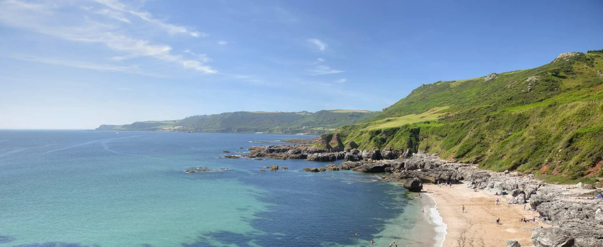 A scenic view of a rocky beach along a green hillside with clear blue ocean and a bright sky.