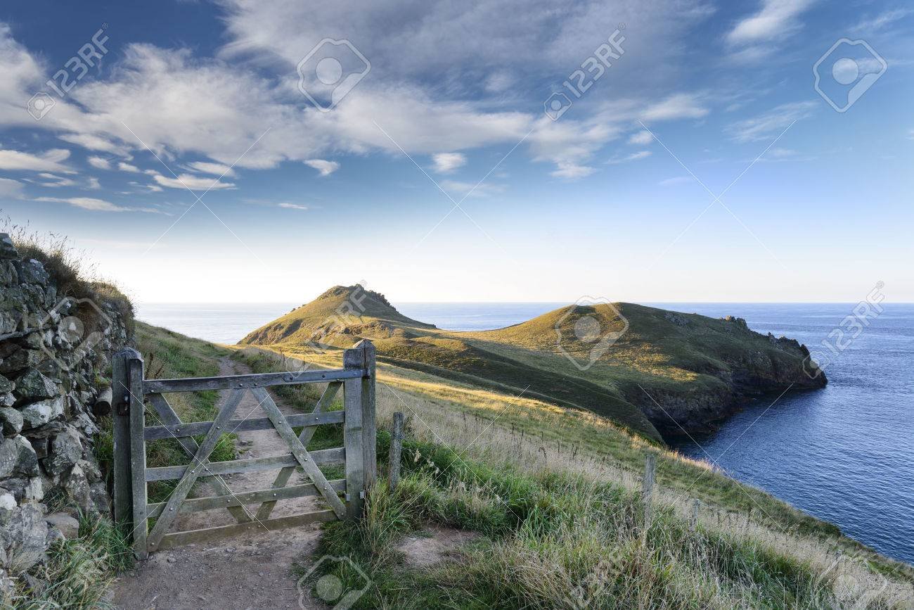 A scenic coastal landscape with a dirt path and wooden gate leading to grassy hills and the ocean in the distance, under a partly cloudy sky.