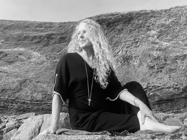 Woman with curly hair sitting on rocks outdoors, looking to the side, wearing a black dress and cross necklace.