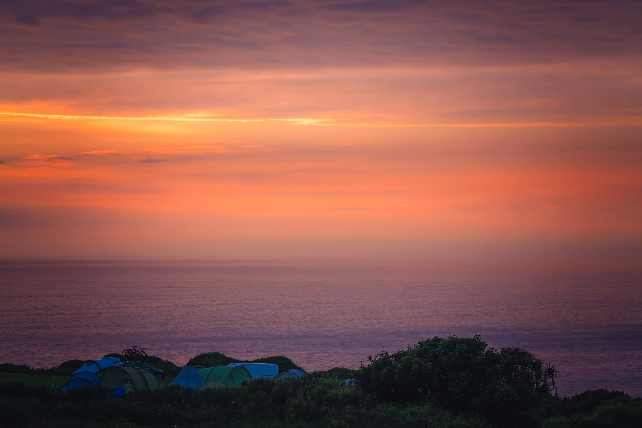 Campground tents set up near the coastline at sunset, with a colorful sky and calm ocean in the background, experience the tranquility at Eden Meadows.