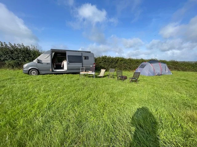 A camping scene with a gray van, a dome tent, and outdoor chairs on a grassy field under a partly cloudy sky.