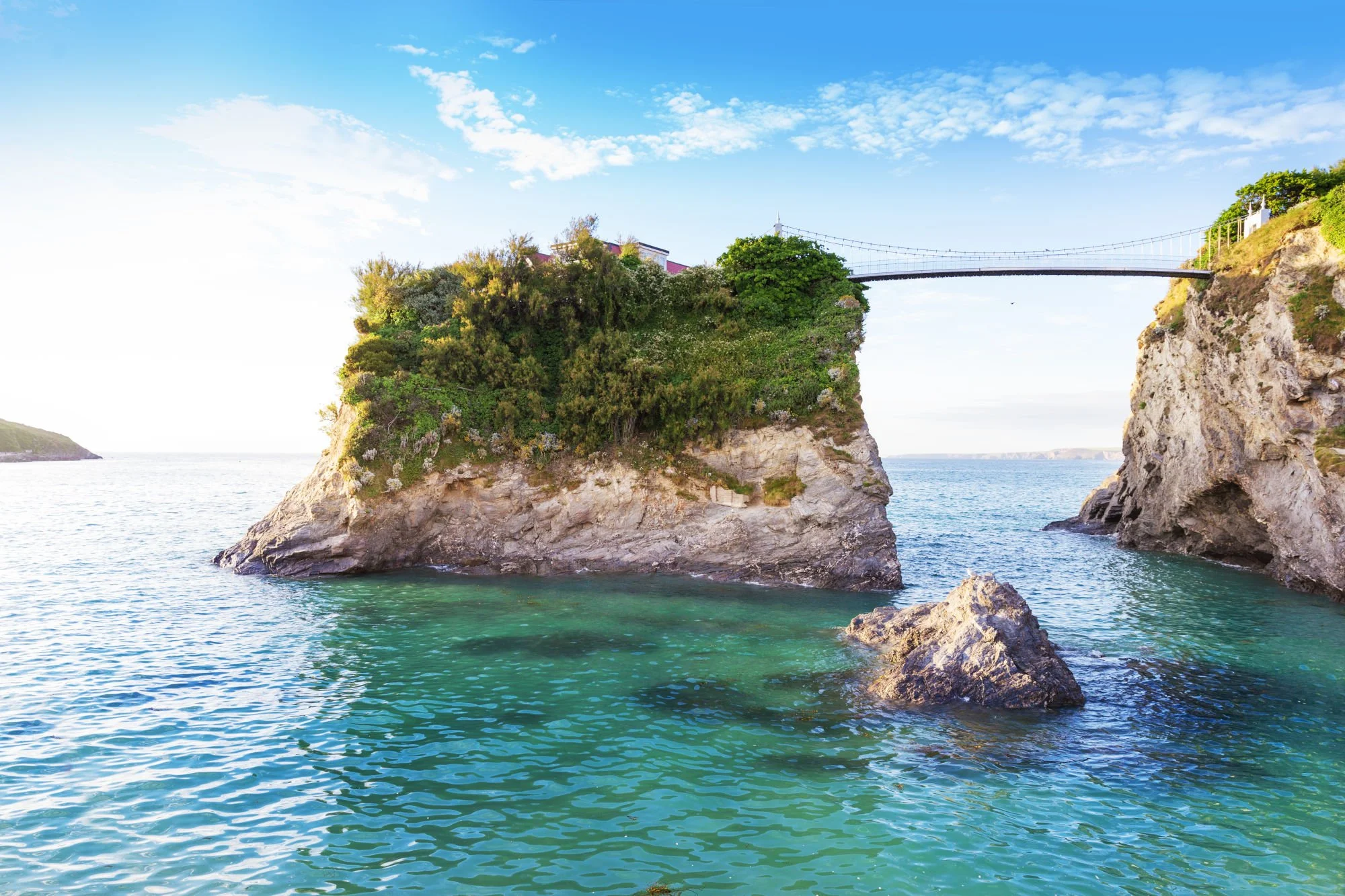 Cliff with lush greenery at the ocean, with a wooden bridge connecting the top of the cliff to another landmass, and a bright blue sky with some clouds.
