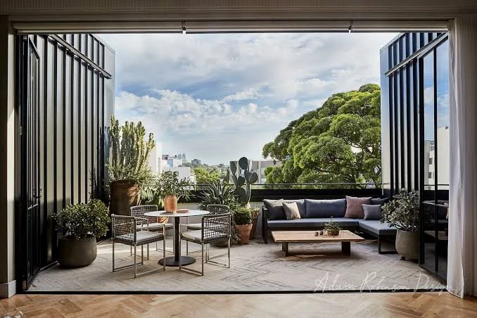 Modern balcony with outdoor furniture, potted plants, and a view of trees and city buildings under a partly cloudy sky.