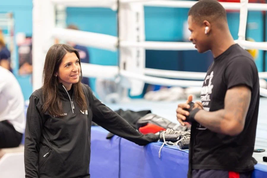A woman in a black athletic jacket speaking with a young man in a gym or boxing ring area.