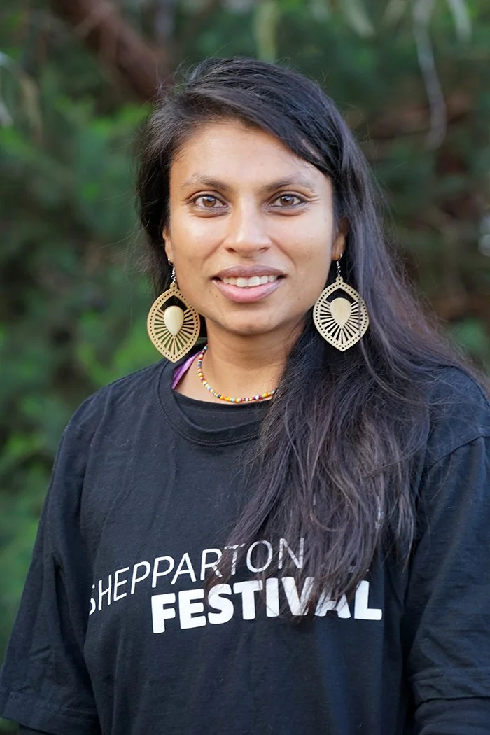 A woman with a black t-shirt and gold earrings smiles , she is standing in front of a group of trees
