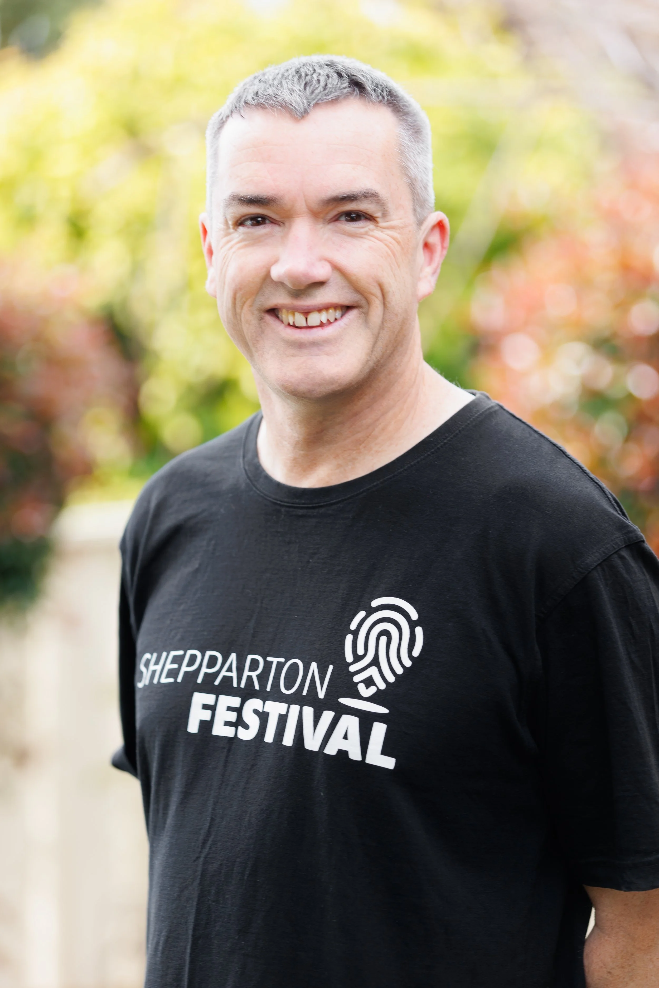 A man with a black t-shirt smiles, he is standing in front of a group of trees