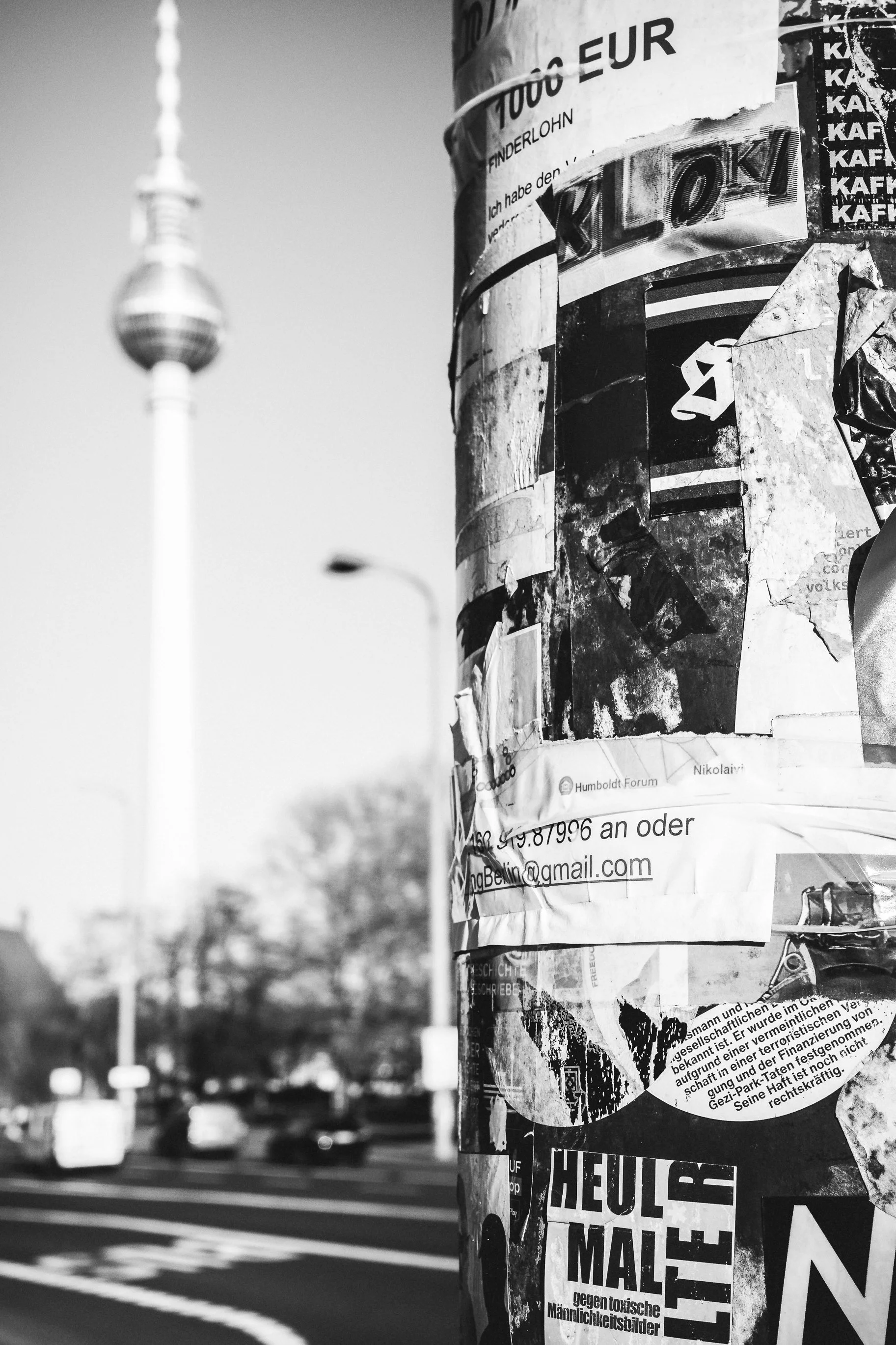 Słup ogłoszeniowy i wieża telewizyjna w Berlinie - czarno-biała fotografia uliczna z architekturą miasta w tle.