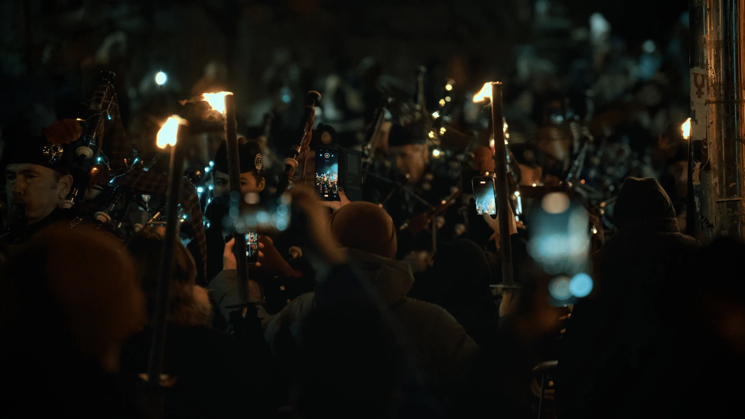 People hold up their phones to film a Scottish marching band walking along the procession route for Edinburgh's 2025 Hogmanay celebration.