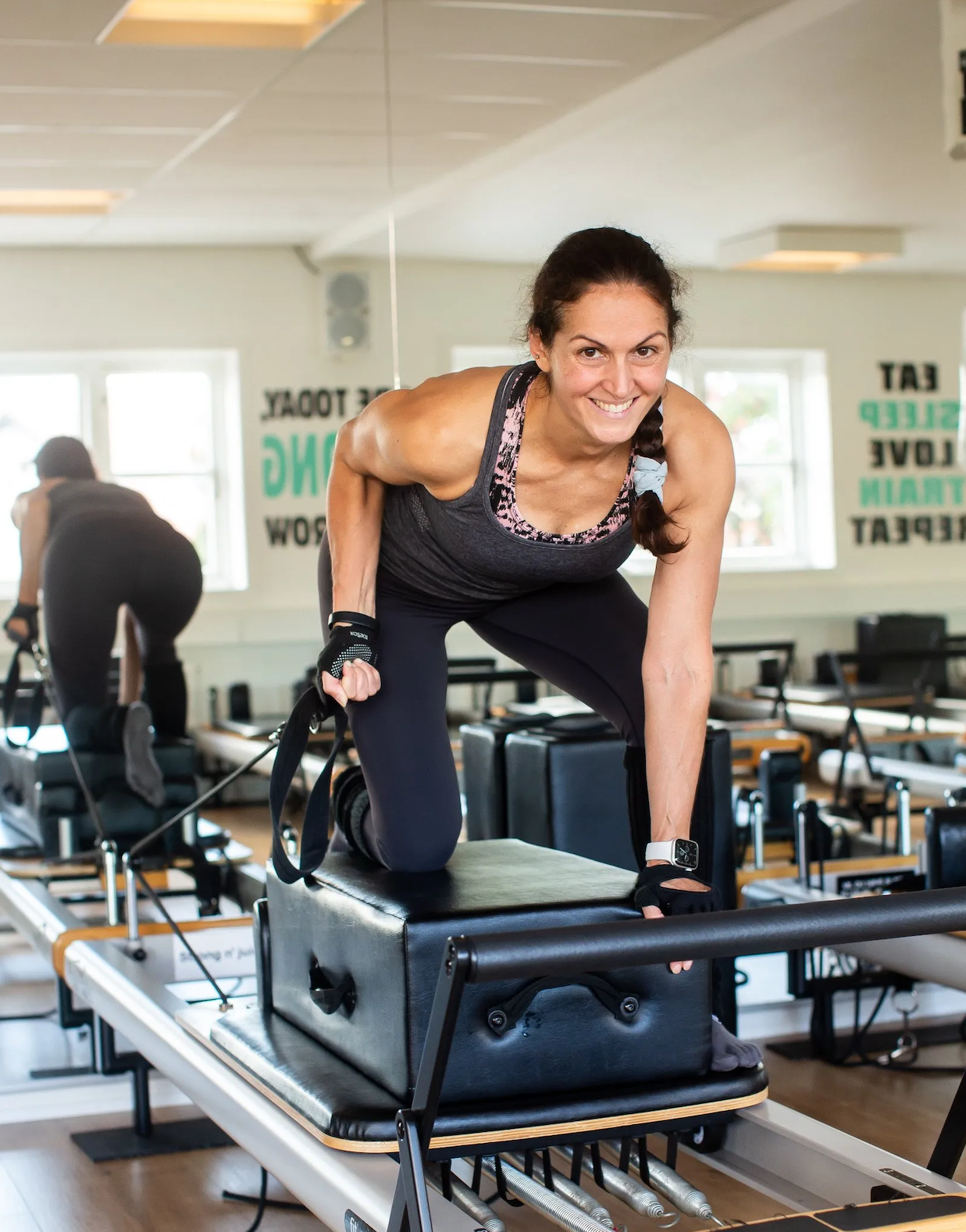 Woman smiling during exercise on Pilates reformer machine in fitness studio, with another person in the background