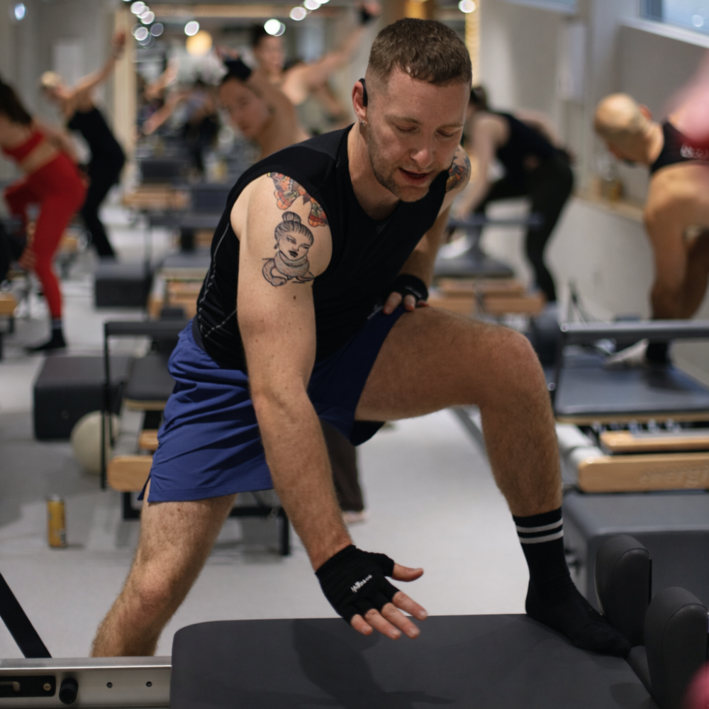 A man with a shaved head, white beard, wearing a white tank top, beige pants, and gray hoodie in a gym, training with a microphone headset on, and people exercising on workout benches in the background.
