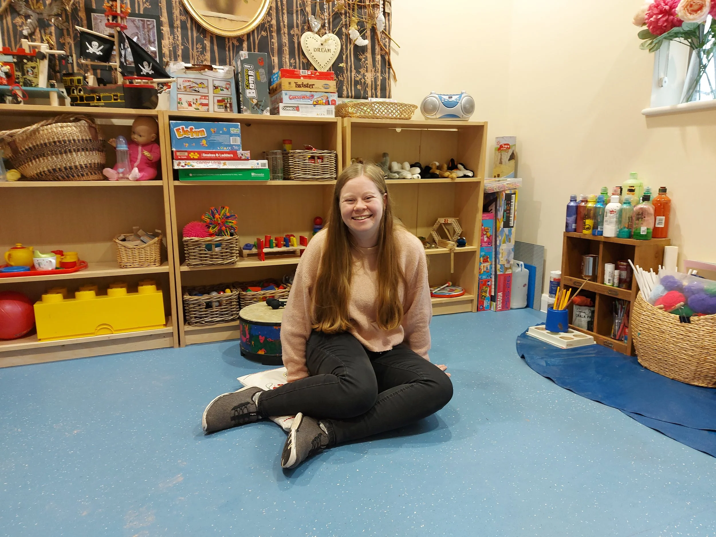 Young woman sitting on the blue floor of a playroom, smiling at the camera. Behind her are shelves filled with toys, books, and art supplies.