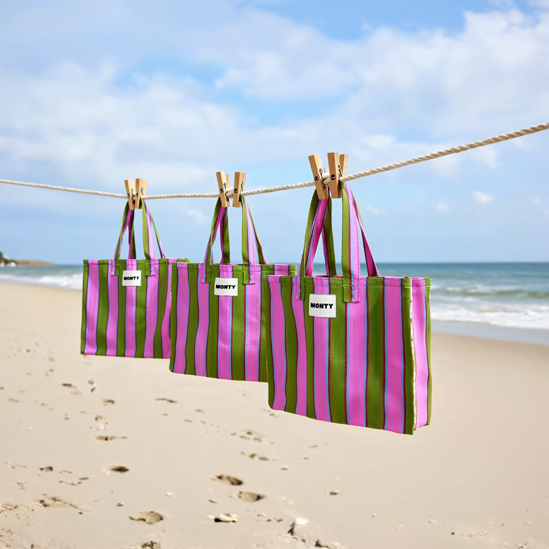 Three pink and green striped tote bags with labels reading 'Monty' hanging on a clothesline at the beach.