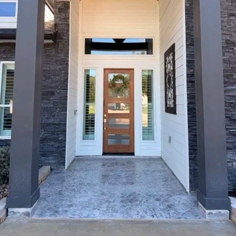 Front porch of a modern house with a wooden door, wreath, sidelight windows, and a transom window above, flanked by stone and white siding.