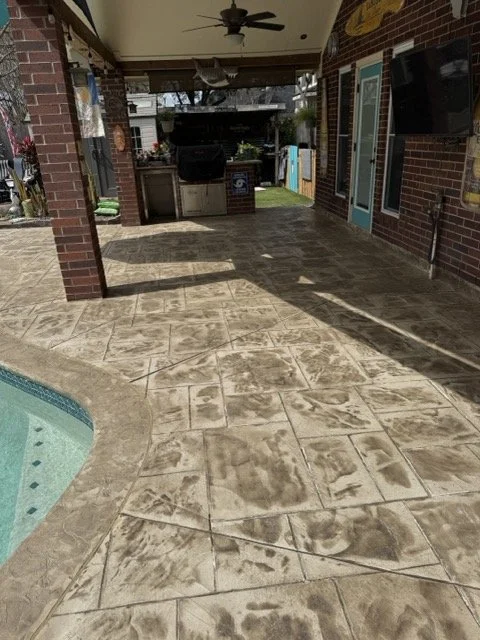 Covered outdoor patio area with brick columns, ceiling fan, ceiling lights, mounted TV, and a fenced backyard with grass and trees in the background.