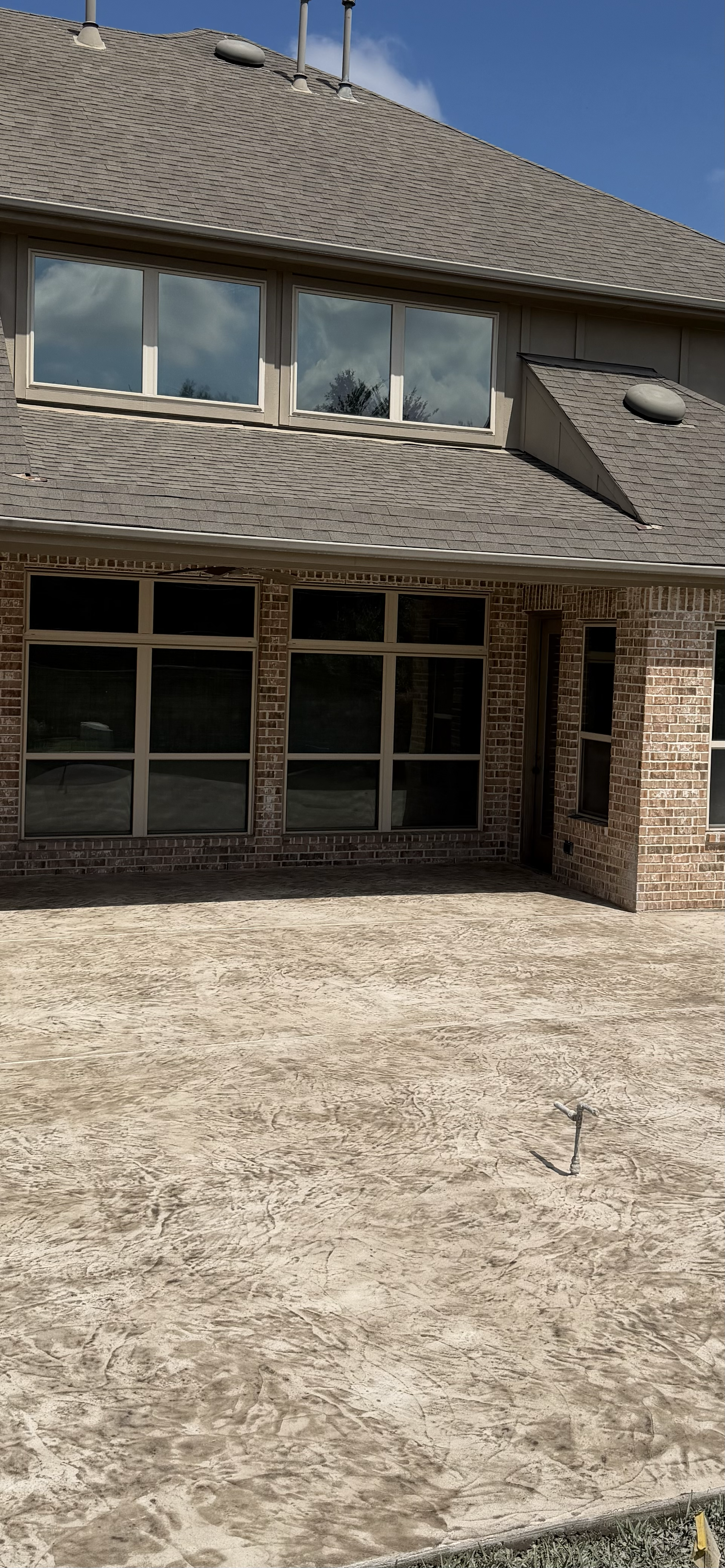 A view of the back of a brick house with large windows, a sloped roof, and a concrete patio or walkway in the foreground. The sky is partly cloudy.
