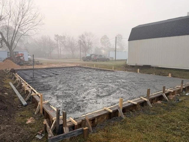 Concrete foundation ready for building, with wooden frame and stakes for formwork, foggy outdoor setting with trees, parked cars, and a white building nearby.