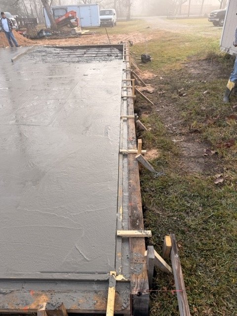 Concrete is being poured and smoothed into a slab on a construction site, with wooden forms and support braces in place along the edges.
