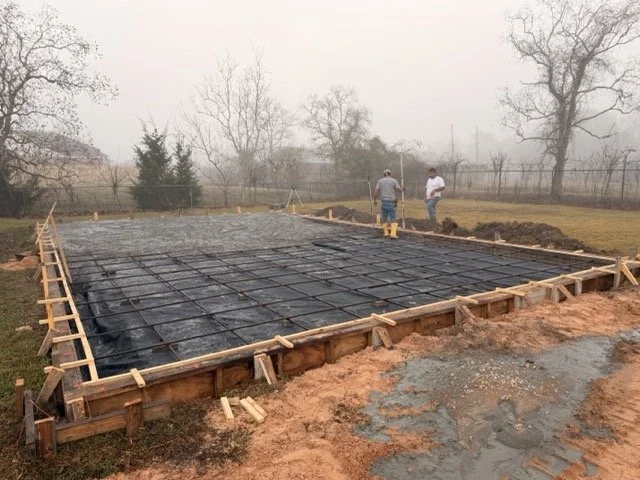 Construction workers preparing a foundation with rebar for a concrete slab in a backyard. The area is fenced with trees in the background during foggy weather.