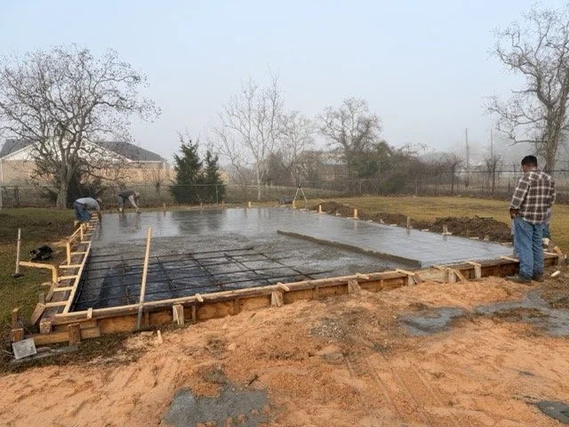 Construction workers pouring concrete for a building foundation on a level wooden form with rebar reinforcement, surrounded by dirt and trees.