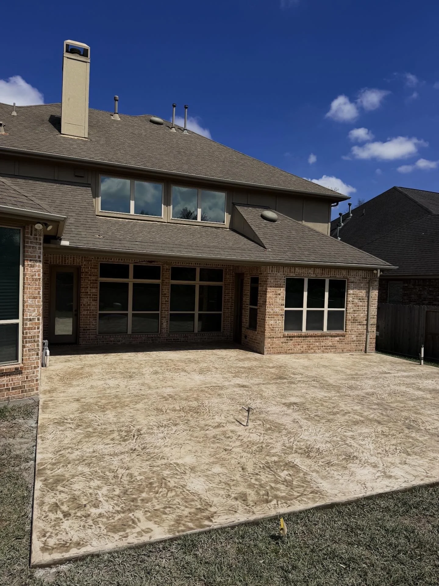 Backyard patio area with stamped concrete flooring, enclosed by brick house walls with large windows, and neighboring house rooftops in the background under a blue sky with some clouds.