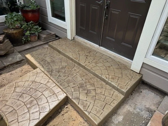 Concrete porch with stamped stone pattern in front of a front door, with potted plants on the side.